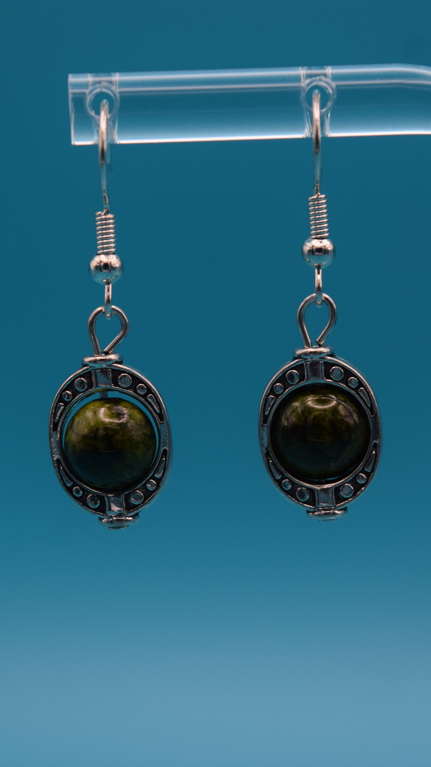 A pair of dangle earrings, each featuring a round, dark green bead set within an ornate, antique silver-colored metal bezel. The metalwork around the stone has a textured, appearance with small decorative dots around the edge. The bead itself is smooth and polished, with variations in its dark green color. This is displayed on a clear stand against a teal background.