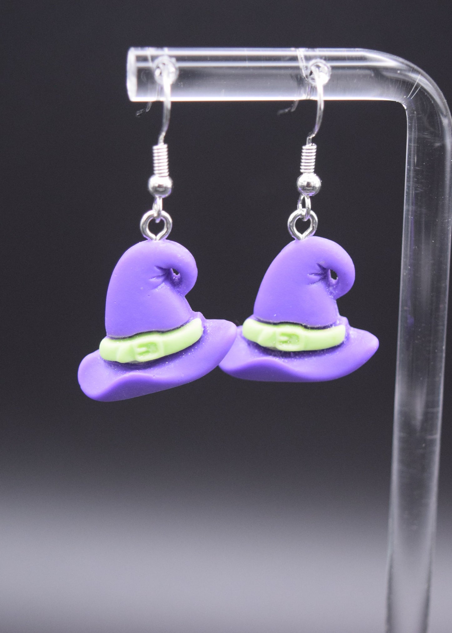Pair of silicone purple witch with a lime green belt hanging 3/4 inches from a silver hook. Displaying on a clear stand against a black background.
