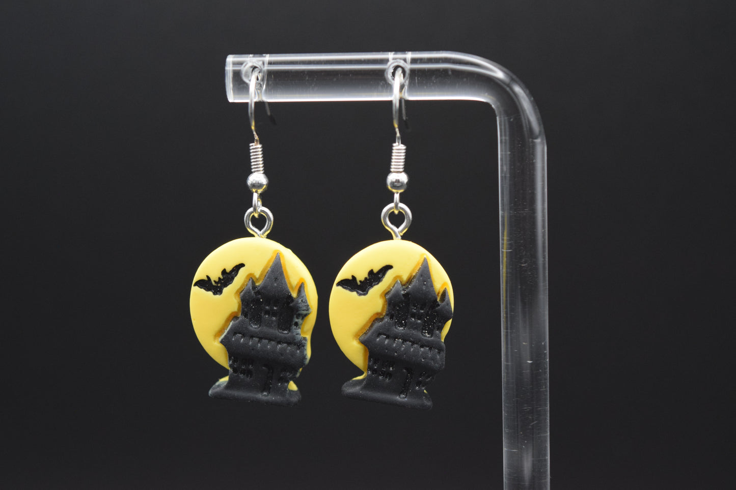 Each earring is a circular design with a bright yellow background, representing a full moon. Silhouetted against this yellow moon are black, elements. In the foreground of each circle is a spooky, multi-storied haunted house, with pointed turrets and a somewhat eerie, stylized appearance. Above the haunted house, flying across the "moon," are small black bats.