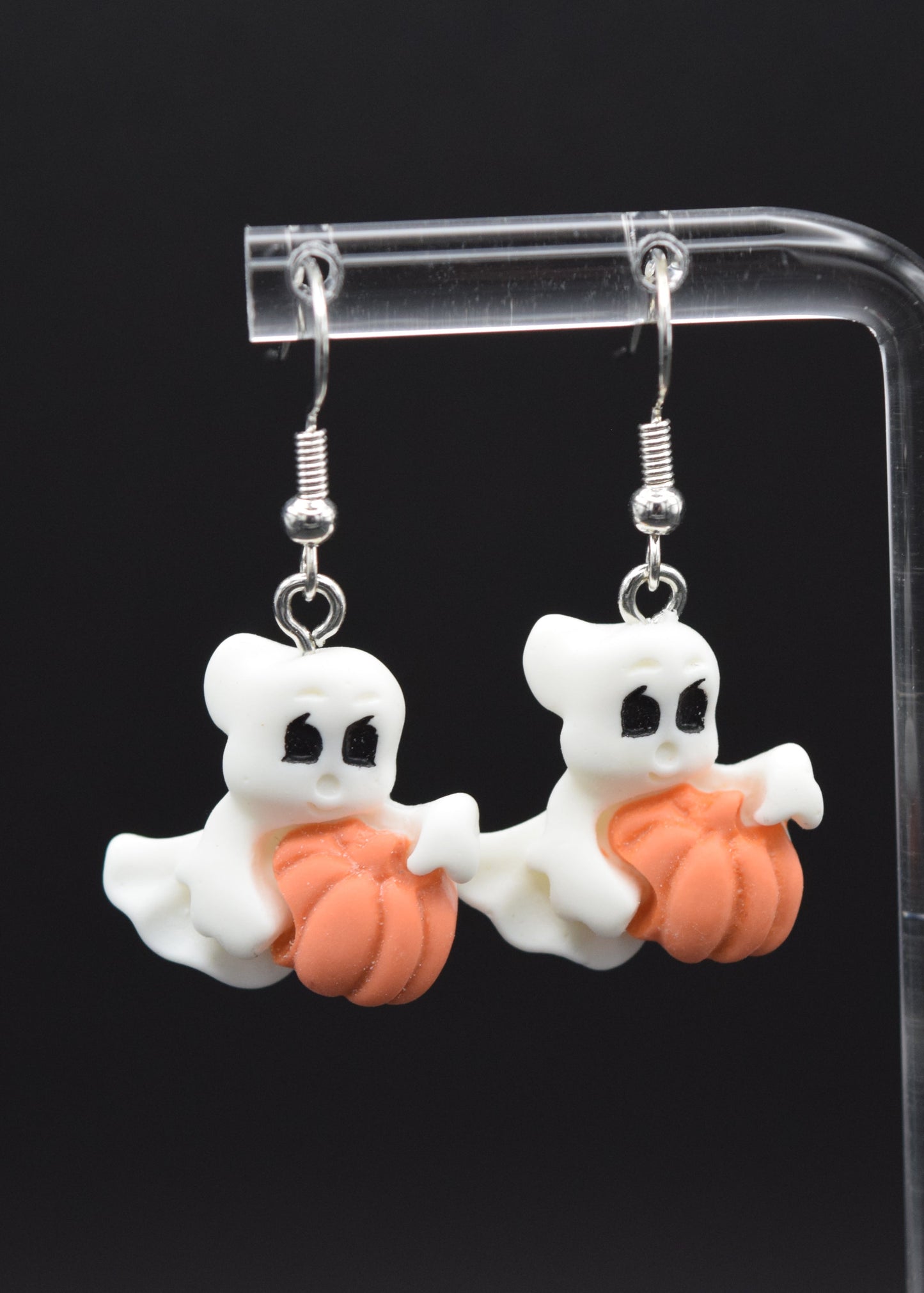 Silicone ghost-shaped earrings with white ghost, with black eyes, holding a orange pumpkins. Displayed on a clear stand against a black background