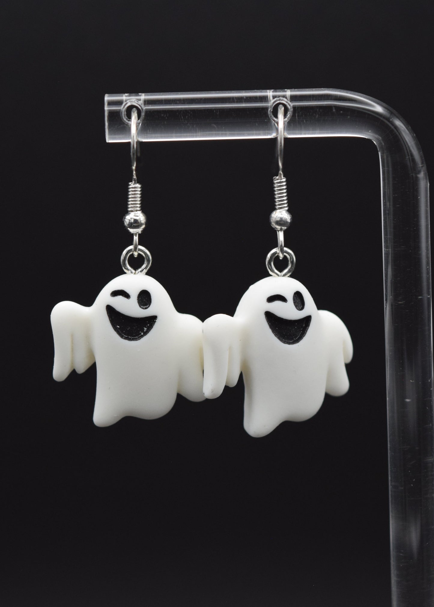 Silicone ghost-shaped earrings with a playful winking expression displayed on a clear stand against a black background