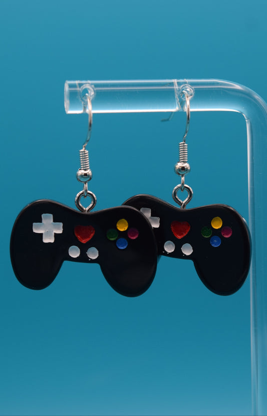 These are a playful and charming pair of dangle game controller earrings, made of black colored epoxy, painted and topped with a clear coat. They're displayed hanging side-by-side on a clear acrylic stand, set against a vibrant, solid teal background.