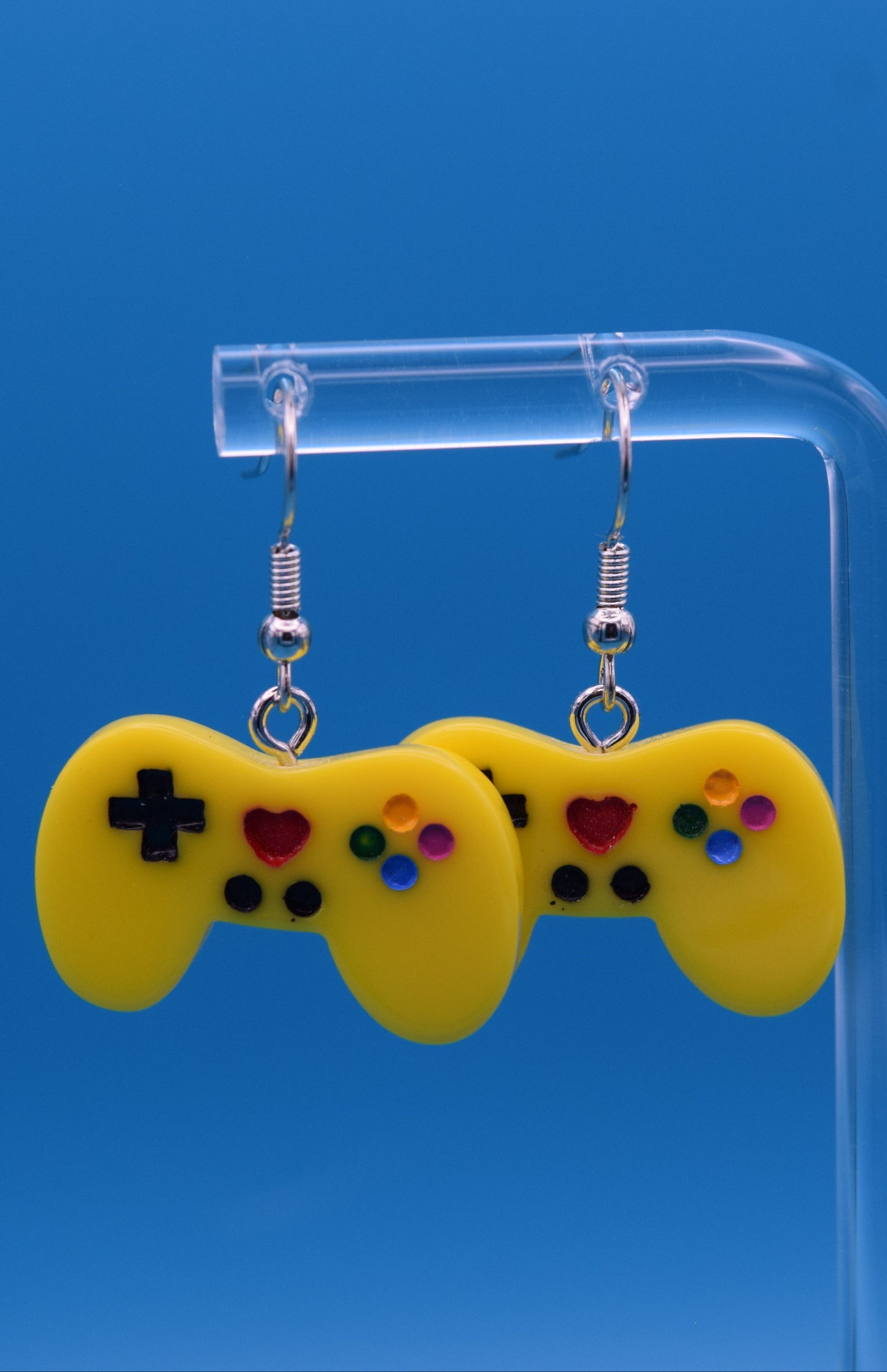 These are a playful and charming pair of dangle game controller earrings, made of colored yellow epoxy, painted and topped with a clear coat. They're displayed hanging side-by-side on a clear acrylic stand, set against a vibrant, solid teal background.