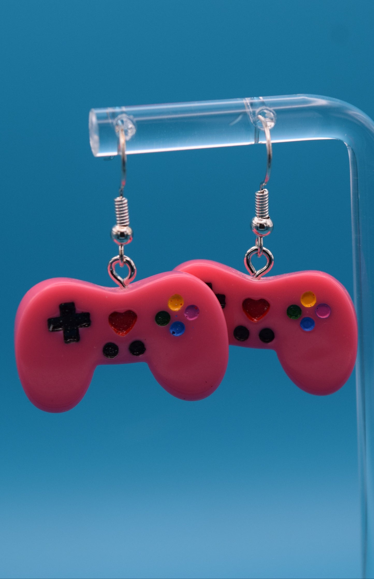These are a playful and charming pair of dangle game controller earrings, made of colored pink epoxy, painted and topped with a clear coat. They're displayed hanging side-by-side on a clear acrylic stand, set against a vibrant, solid teal background.