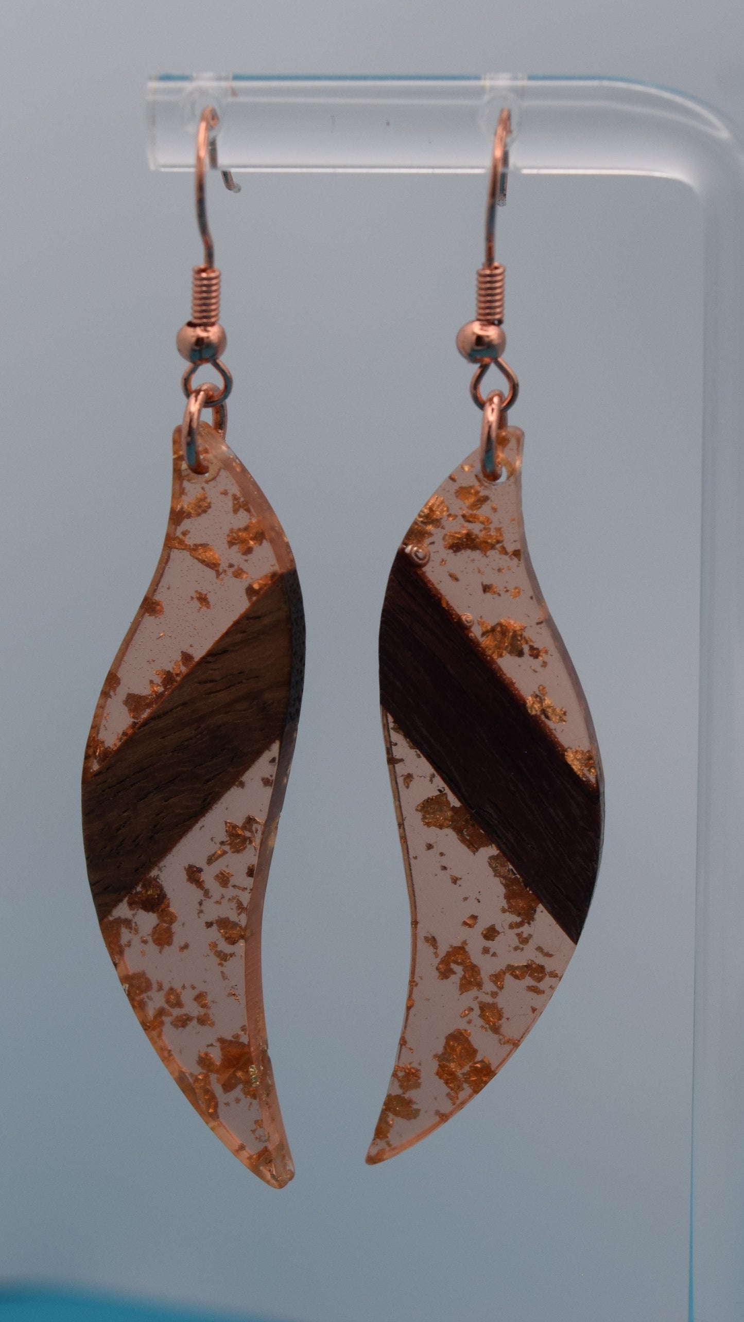 The earring is a translucent orange resin. This resin has a smooth, polished finish, allowing some light to pass through, giving it a luminous quality. Embedded within the resin are numerous small, irregular rose gold flakes with a walnut color piece of wood set in the middle of the earring. These flakes are scattered randomly, catching the light and adding a subtle, delicate shimmer and texture to the surface.