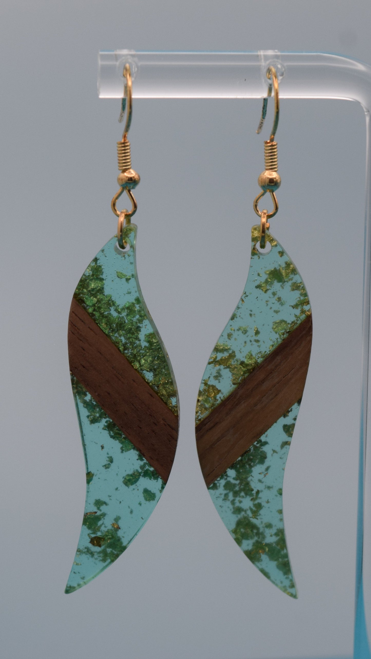 The earring is a translucent light teal resin. This resin has a smooth, polished finish, allowing some light to pass through, giving it a luminous quality. Embedded within the resin are numerous small, irregular gold flakes with a walnut color piece of wood set in the middle of the earring. These flakes are scattered randomly, catching the light and adding a subtle, delicate shimmer and texture to the surface.