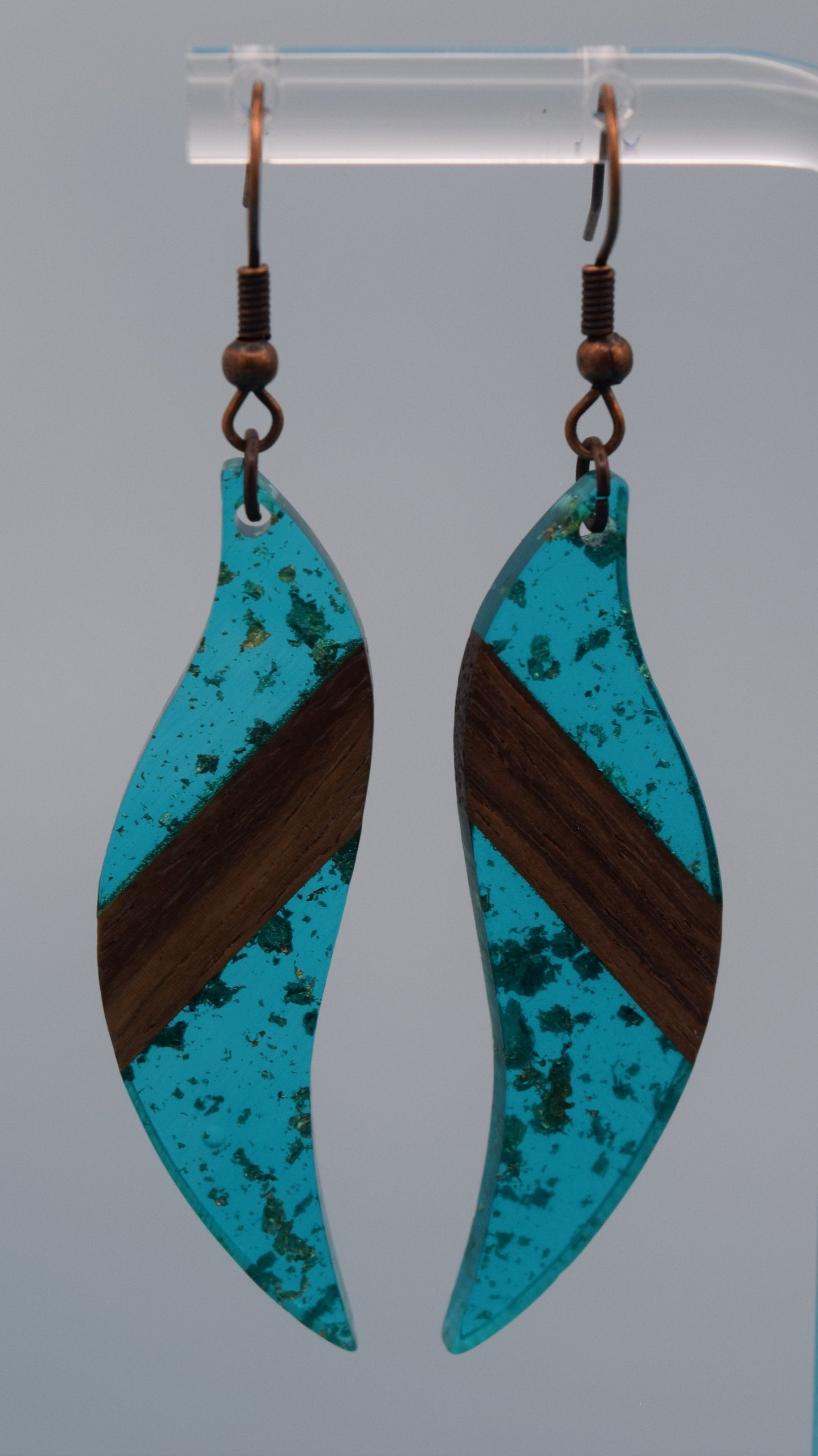 The earring is a translucent turquoise resin. This resin has a smooth, polished finish, allowing some light to pass through, giving it a luminous quality. Embedded within the resin are numerous small, irregular gold flakes with a walnut color piece of wood set in the middle of the earring. These flakes are scattered randomly, catching the light and adding a subtle, delicate shimmer and texture to the surface.