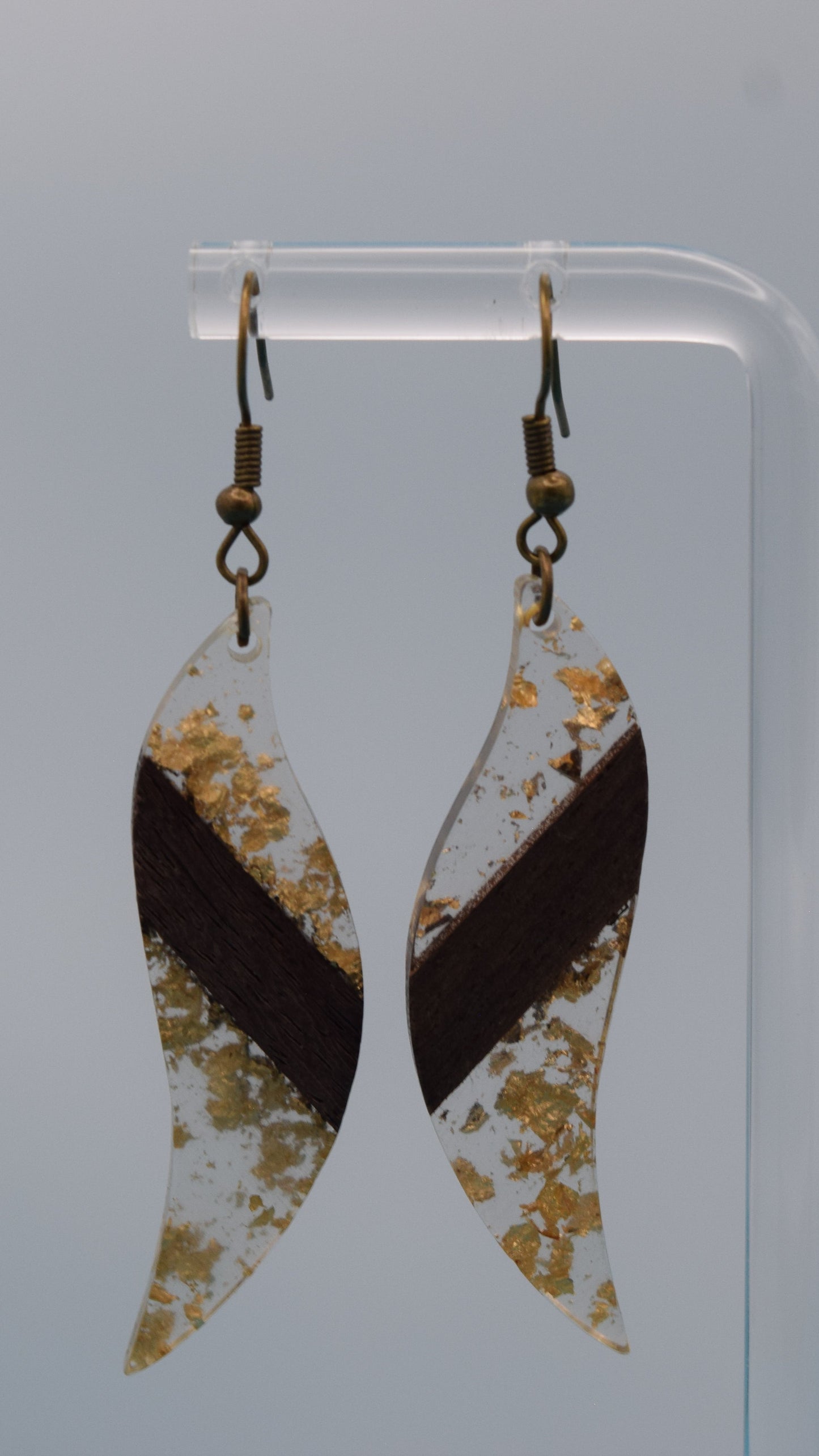 The earring is a translucent resin. This resin has a smooth, polished finish, allowing some light to pass through, giving it a luminous quality. Embedded within the resin are numerous small, irregular gold flakes with a walnut color piece of wood set in the middle of the earring. These flakes are scattered randomly, catching the light and adding a subtle, delicate shimmer and texture to the surface.
