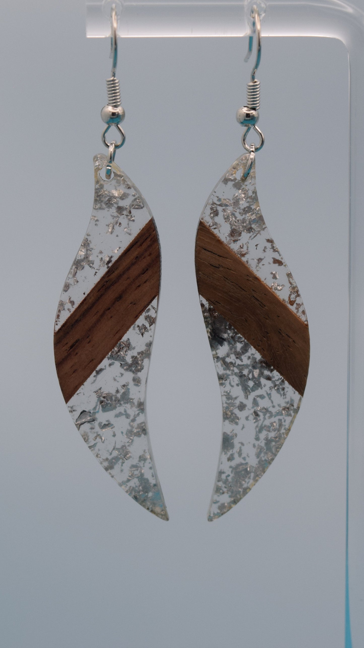 The earring is a translucent resin. This resin has a smooth, polished finish, allowing some light to pass through, giving it a luminous quality. Embedded within the resin are numerous small, irregular silver flakes with a walnut color piece of wood set in the middle of the earring. These flakes are scattered randomly, catching the light and adding a subtle, delicate shimmer and texture to the surface.