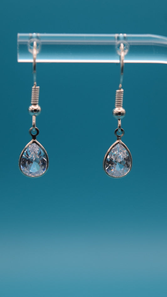 Teardrop-shaped dangling earrings hanging at 1/2 inch long featuring silver teardrop edge with a clear crystal incased at the center. Displayed on a clear stand against a teal background.