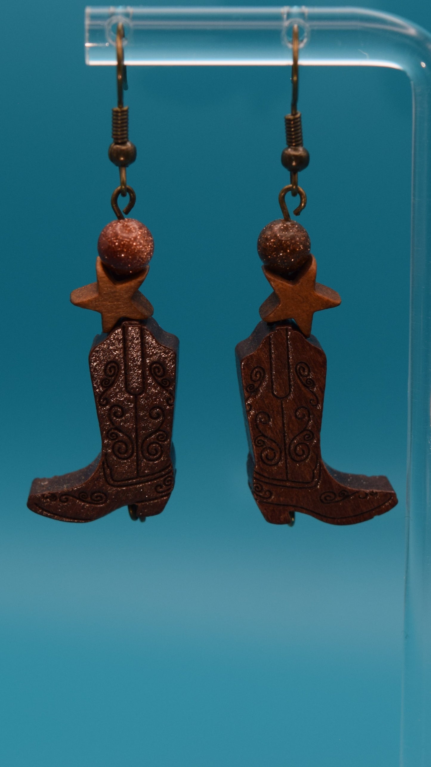 Wooden cowboy boot with a wooden star and a bronze sparkle bead earrings. Displayed on a clear stand against a teal background.