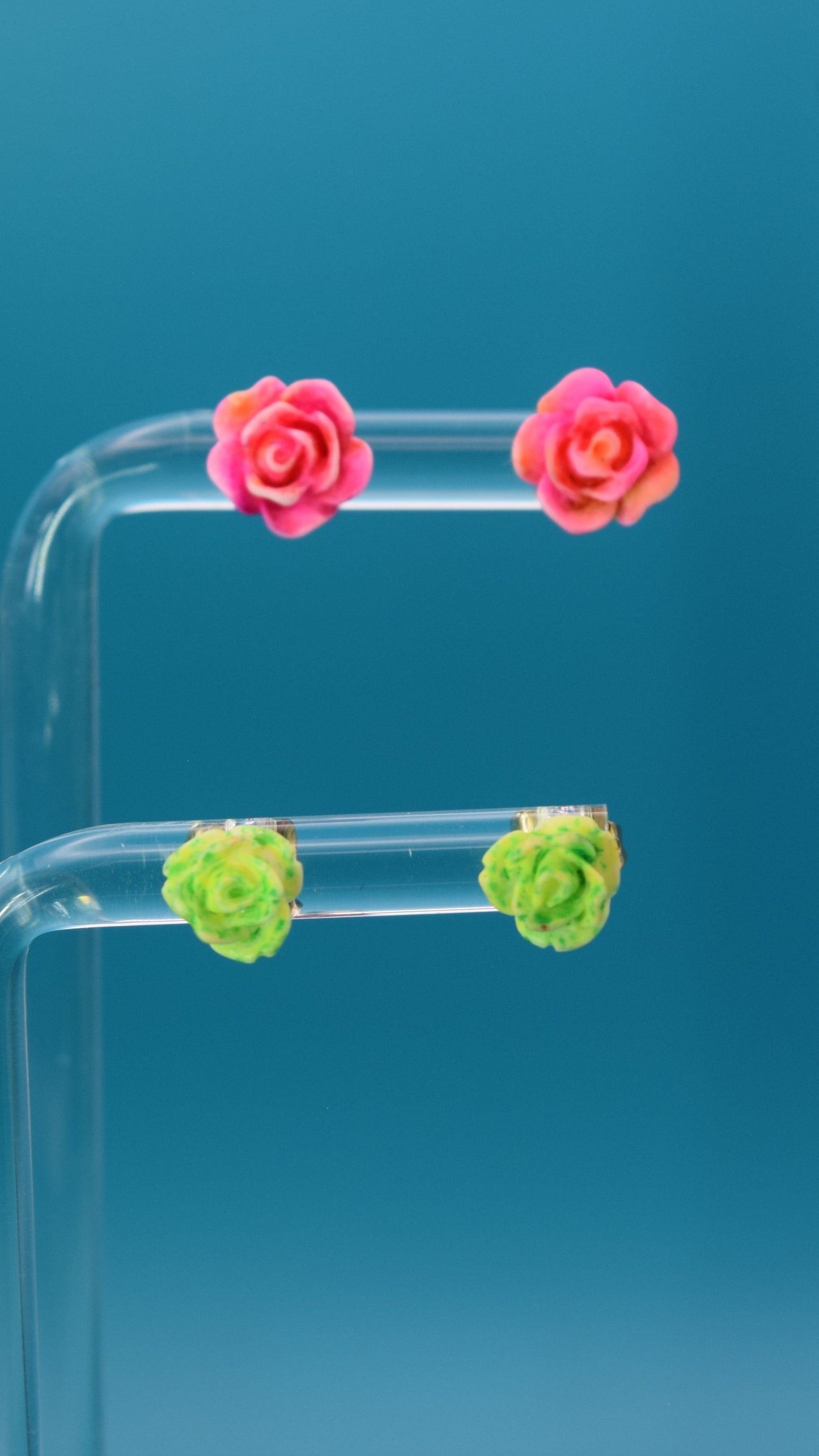 Hot pink and orange tie dyed effect rose-shaped stud earrings, paired with a smaller yellow and green tie dyed effect rose. Displayed on a clear stand against a teal background.