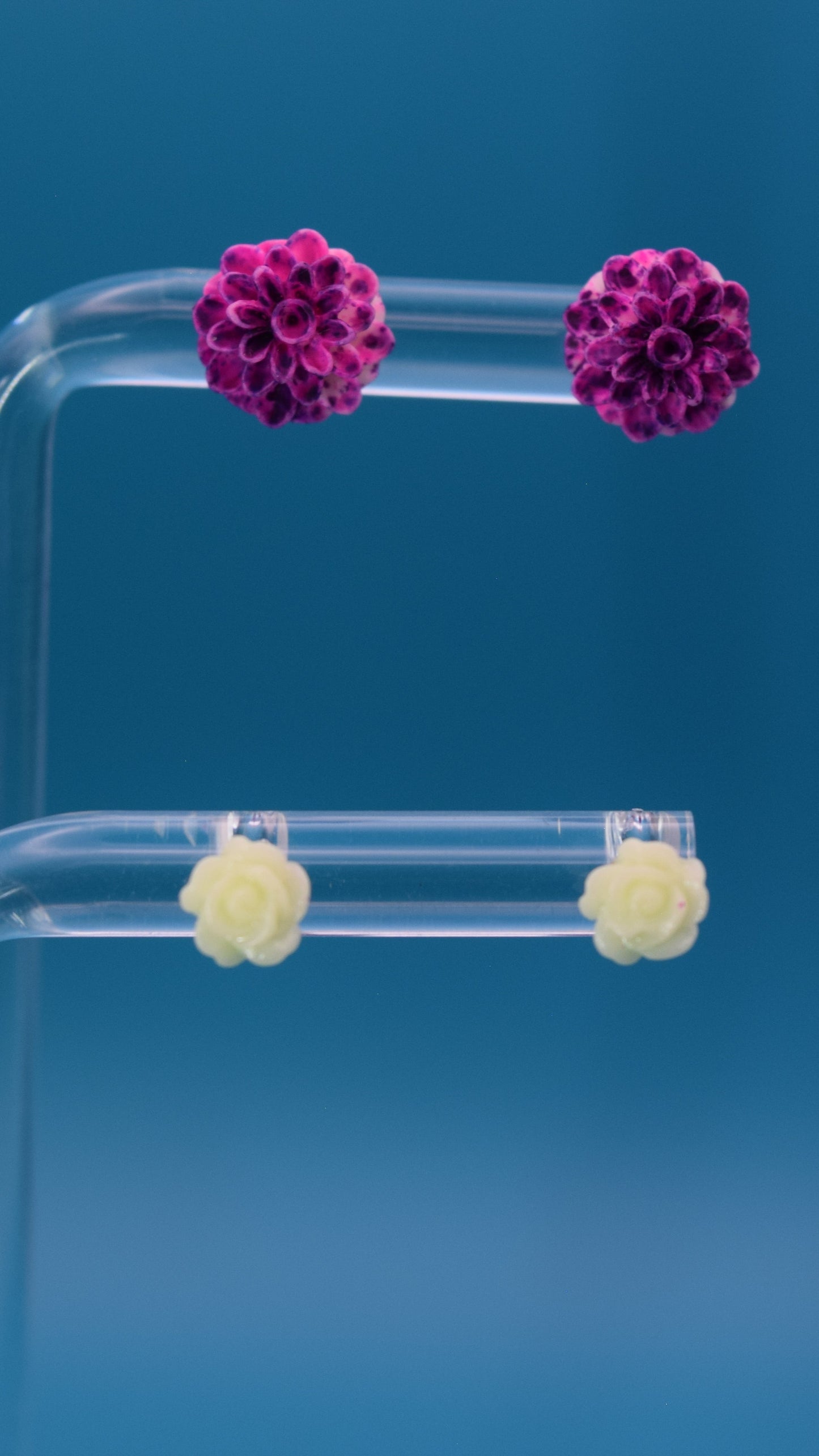 light and dark purple colored dahlia flower-shaped stud earrings, paired with light green almost white colored small rose. Displayed on a clear stand against a teal