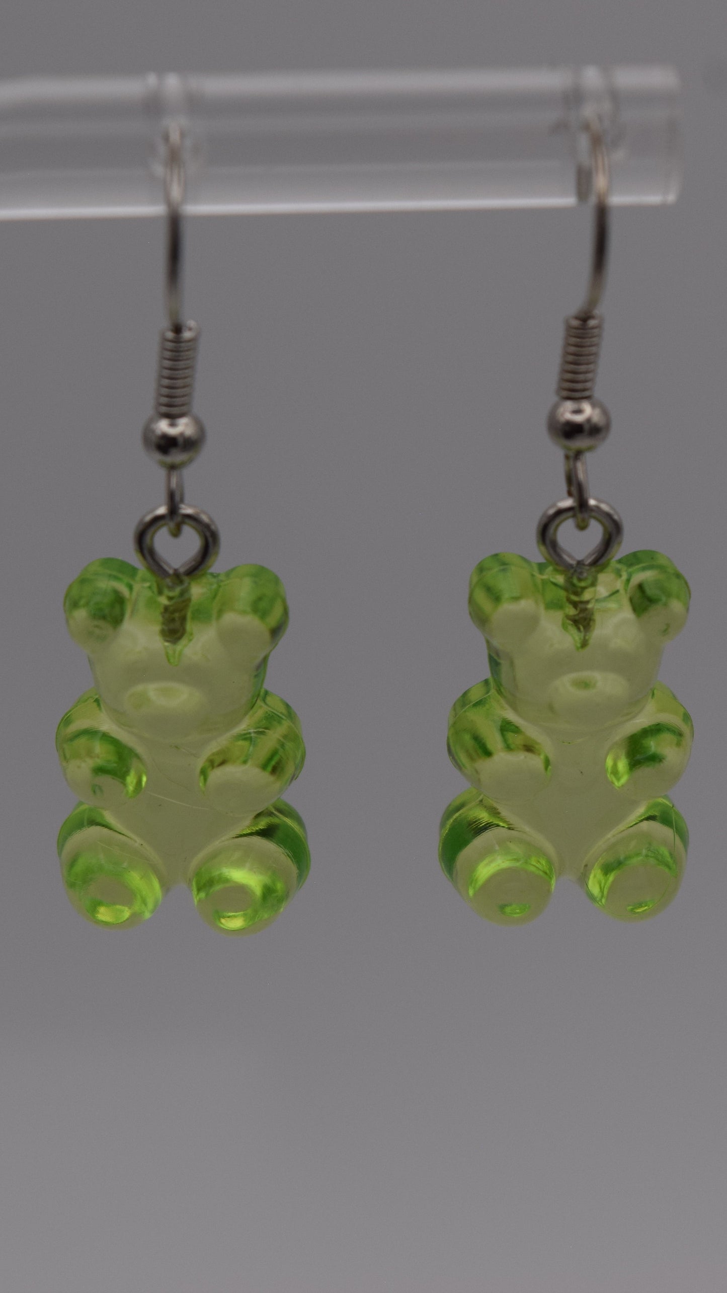 Epoxy 2 inch green gummy bear-shaped earrings with silver earring hook. Displayed on a clear stand against a gray background.