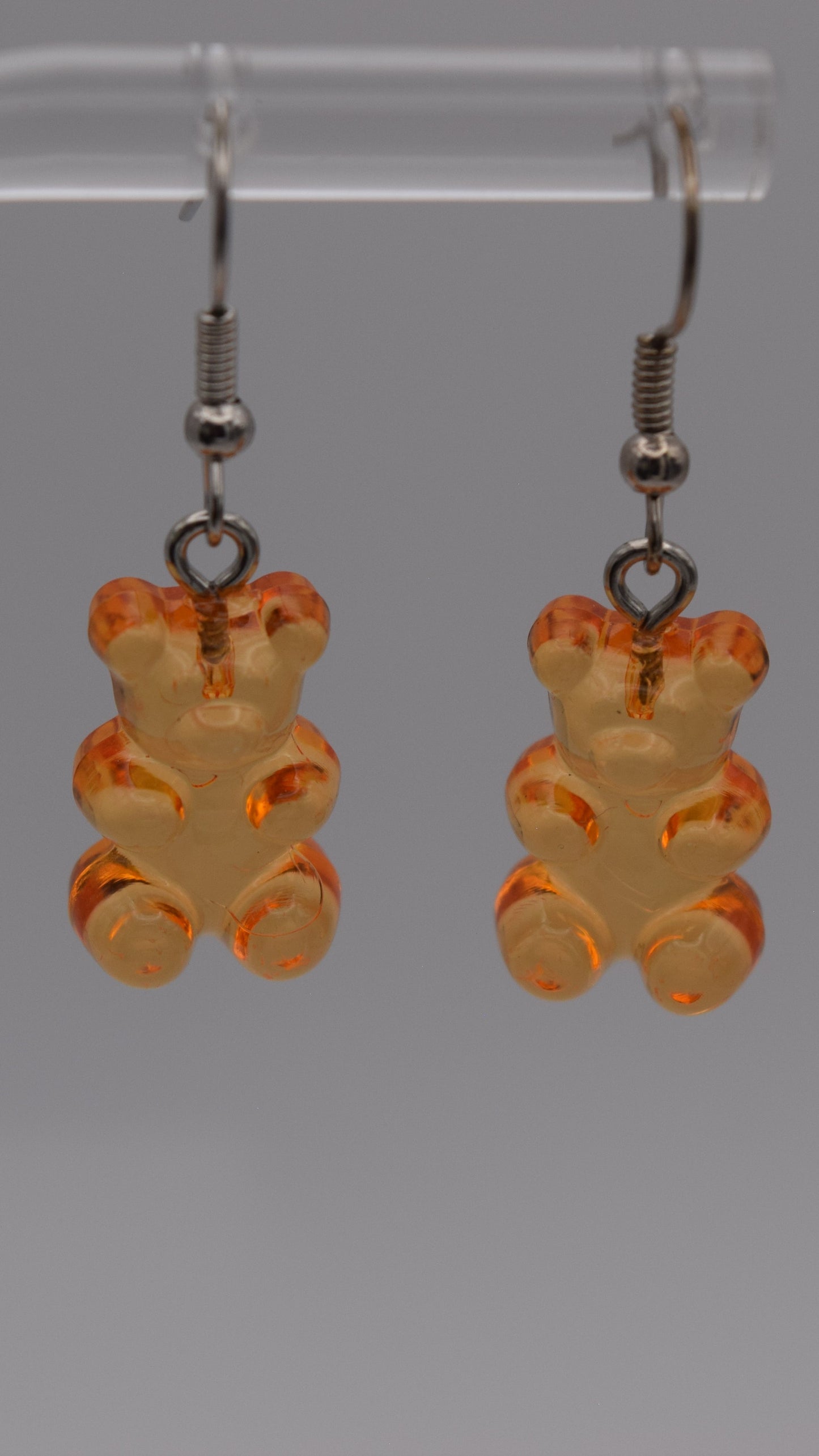 Epoxy 2 inch orange gummy bear-shaped earrings with silver earring hook. Displayed on a clear stand against a gray background.