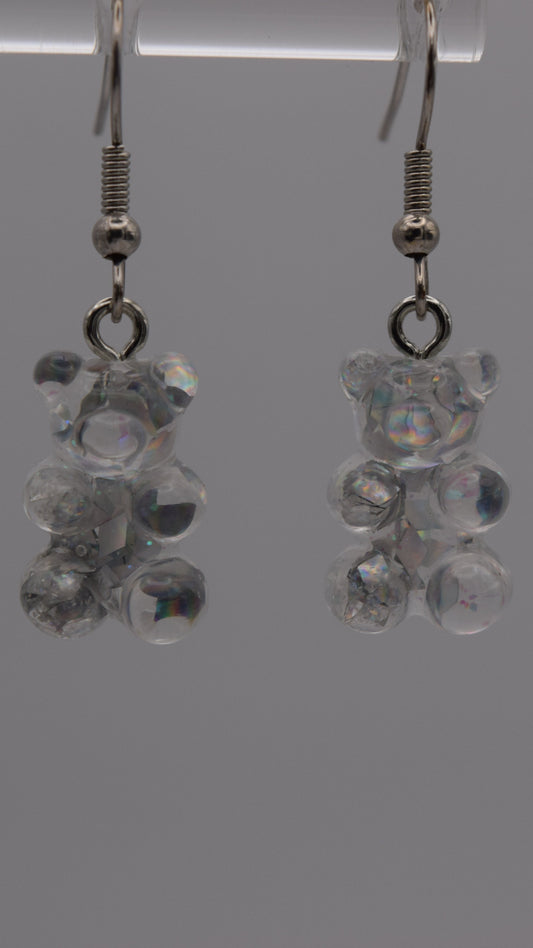 Epoxy 2 inch gummy bear-shaped earrings, clear with iridescent flacks with silver earring hook. Displayed on a clear stand against a gray background.