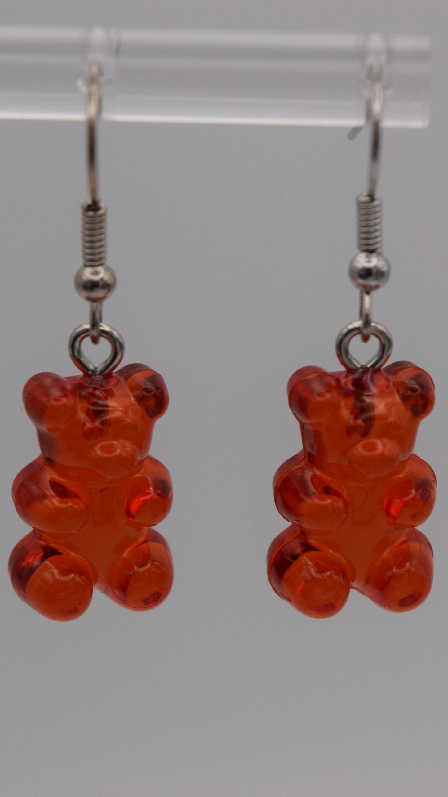 Epoxy 2 inch red gummy bear-shaped earrings with silver earring hook. Displayed on a clear stand against a gray background.