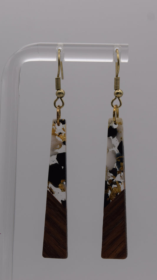 These are a pair of dangle earrings, each earring consists of a long, rectangular drop. The drops are made of two distinct sections. The bottom third of each earring is a solid, dark brown wood, smoothly polished. Above this wooden section, the remaining two-thirds of the earring is made of a clear, glossy resin material. Within this resin, there's a striking abstract design. It's primarily an epoxy mix of clear, white, black, and gold flakes.
