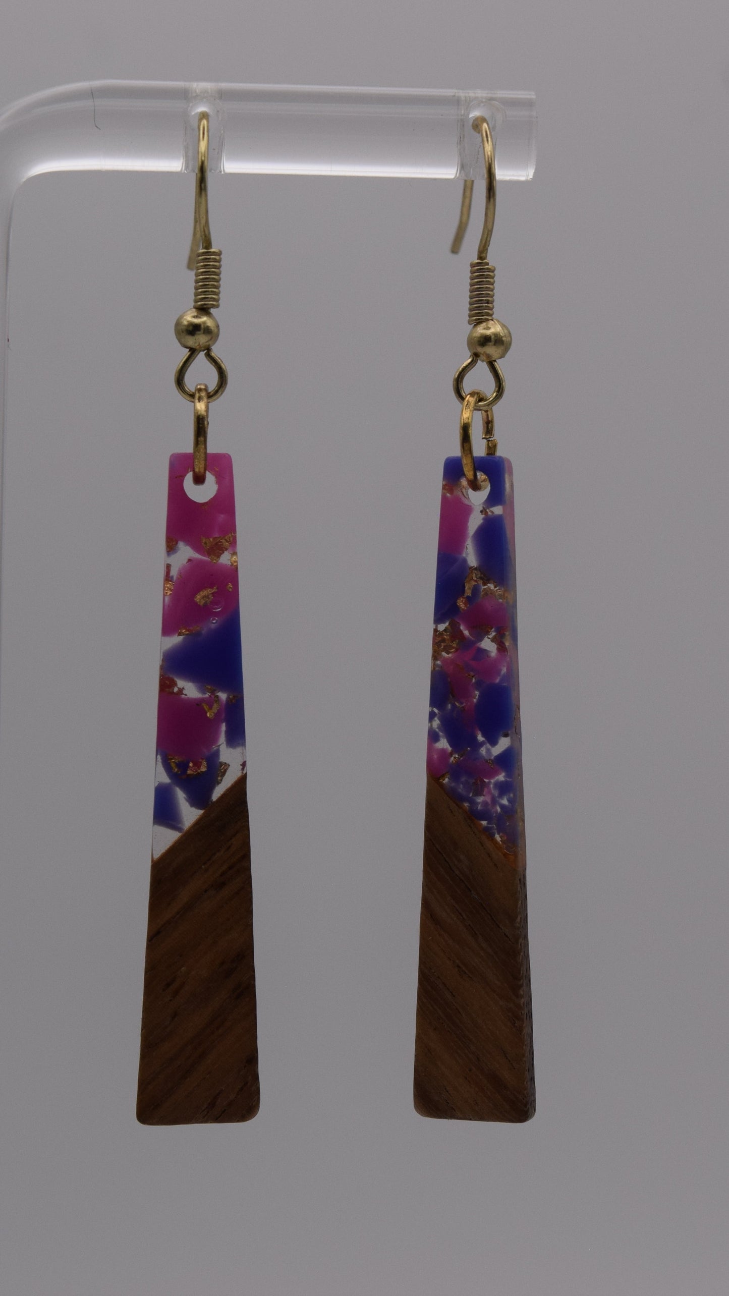 These are a pair of dangle earrings, each earring consists of a long, rectangular drop. The drops are made of two distinct sections. The bottom third of each earring is a solid, dark brown wood, smoothly polished. Above this wooden section, the remaining two-thirds of the earring is made of a clear, glossy resin material. Within this resin, there's a striking abstract design. It's primarily an epoxy mix of clear, pink, purple, and gold flakes.