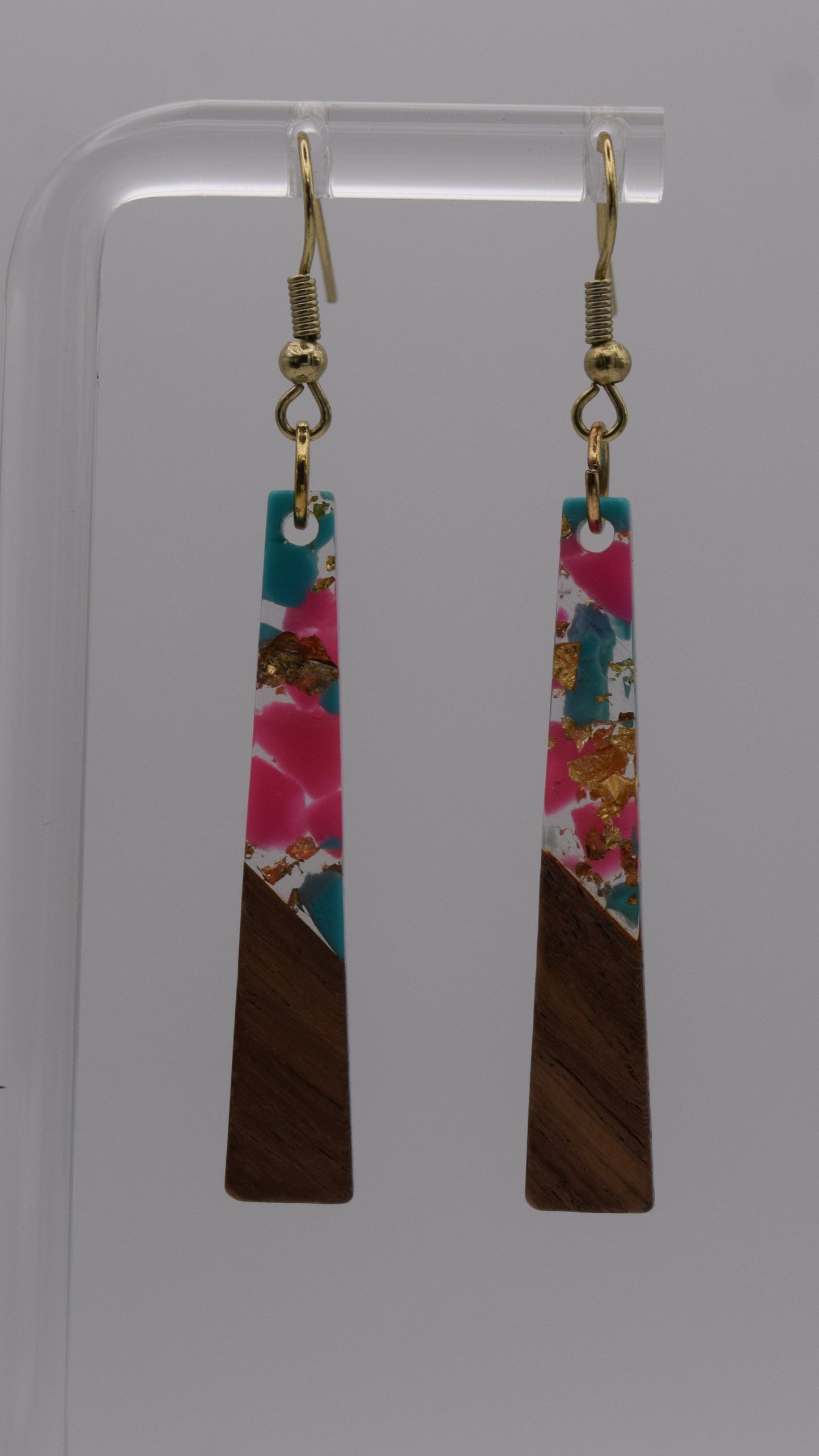 These are a pair of dangle earrings, each earring consists of a long, rectangular drop. The drops are made of two distinct sections. The bottom third of each earring is a solid, dark brown wood, smoothly polished. Above this wooden section, the remaining two-thirds of the earring is made of a clear, glossy resin material. Within this resin, there's a striking abstract design. It's primarily an epoxy mix of clear, teal, pink, and gold flakes.