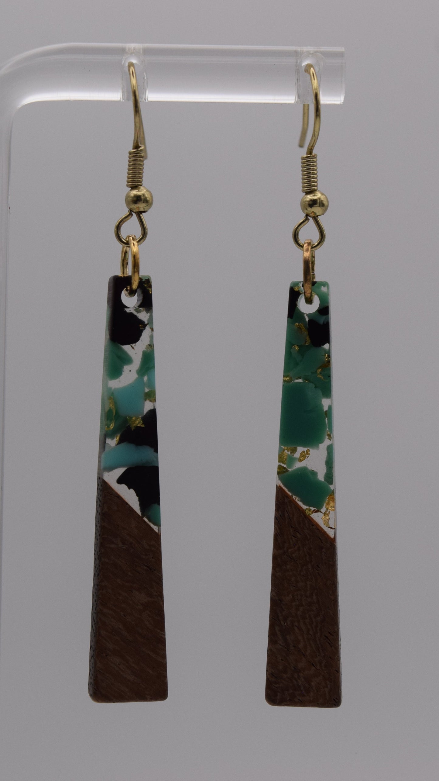 These are a pair of dangle earrings, each earring consists of a long, rectangular drop. The drops are made of two distinct sections. The bottom third of each earring is a solid, dark brown wood, smoothly polished. Above this wooden section, the remaining two-thirds of the earring is made of a clear, glossy resin material. Within this resin, there's a striking abstract design. It's primarily an epoxy mix of clear, teal, black, and gold flakes.
