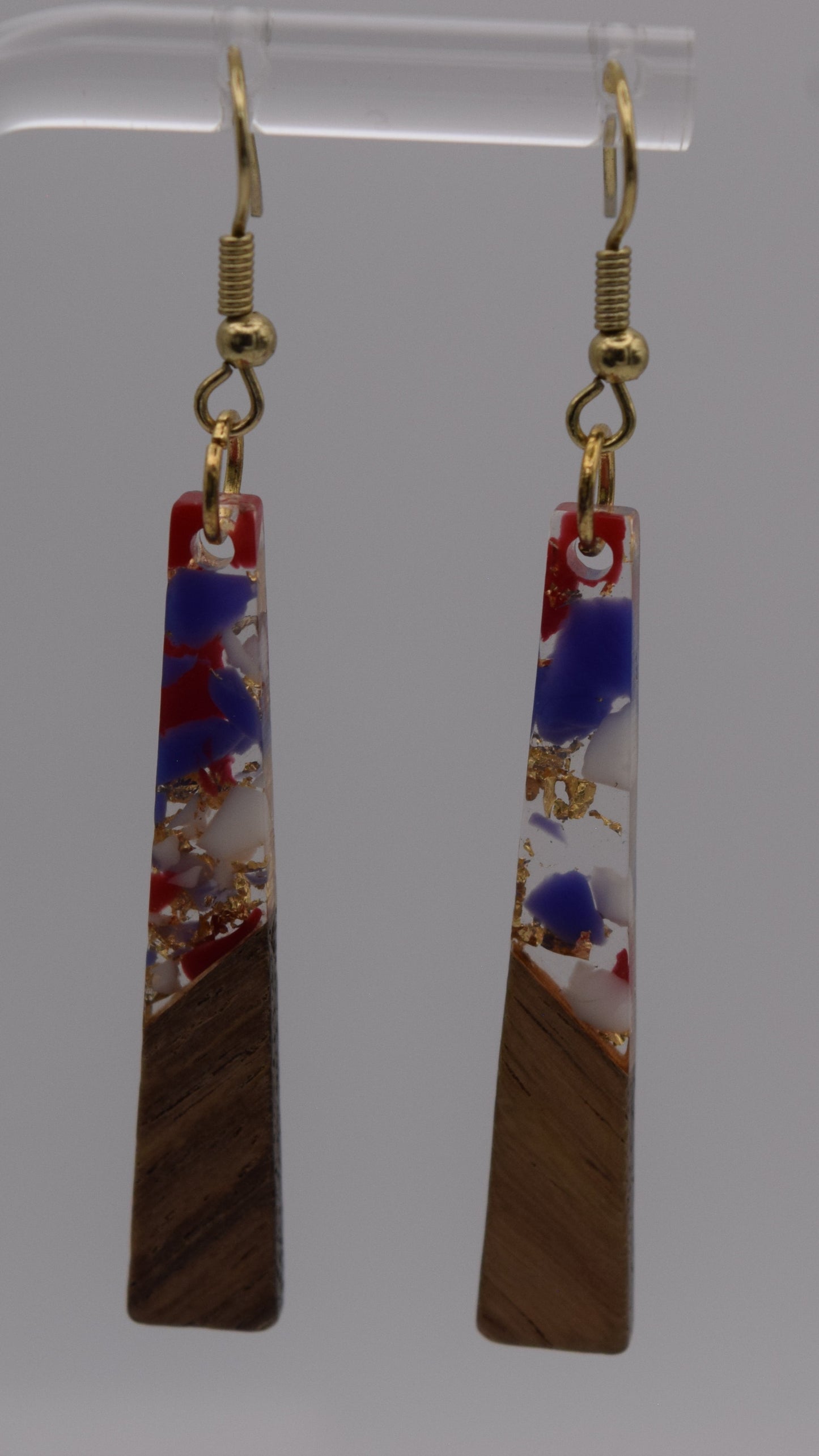 These are a pair of dangle earrings, each earring consists of a long, rectangular drop. The drops are made of two distinct sections. The bottom third of each earring is a solid, dark brown wood, smoothly polished. Above this wooden section, the remaining two-thirds of the earring is made of a clear, glossy resin material. Within this resin, there's a striking abstract design. It's primarily an epoxy mix of clear, red, blue, white and gold flakes.