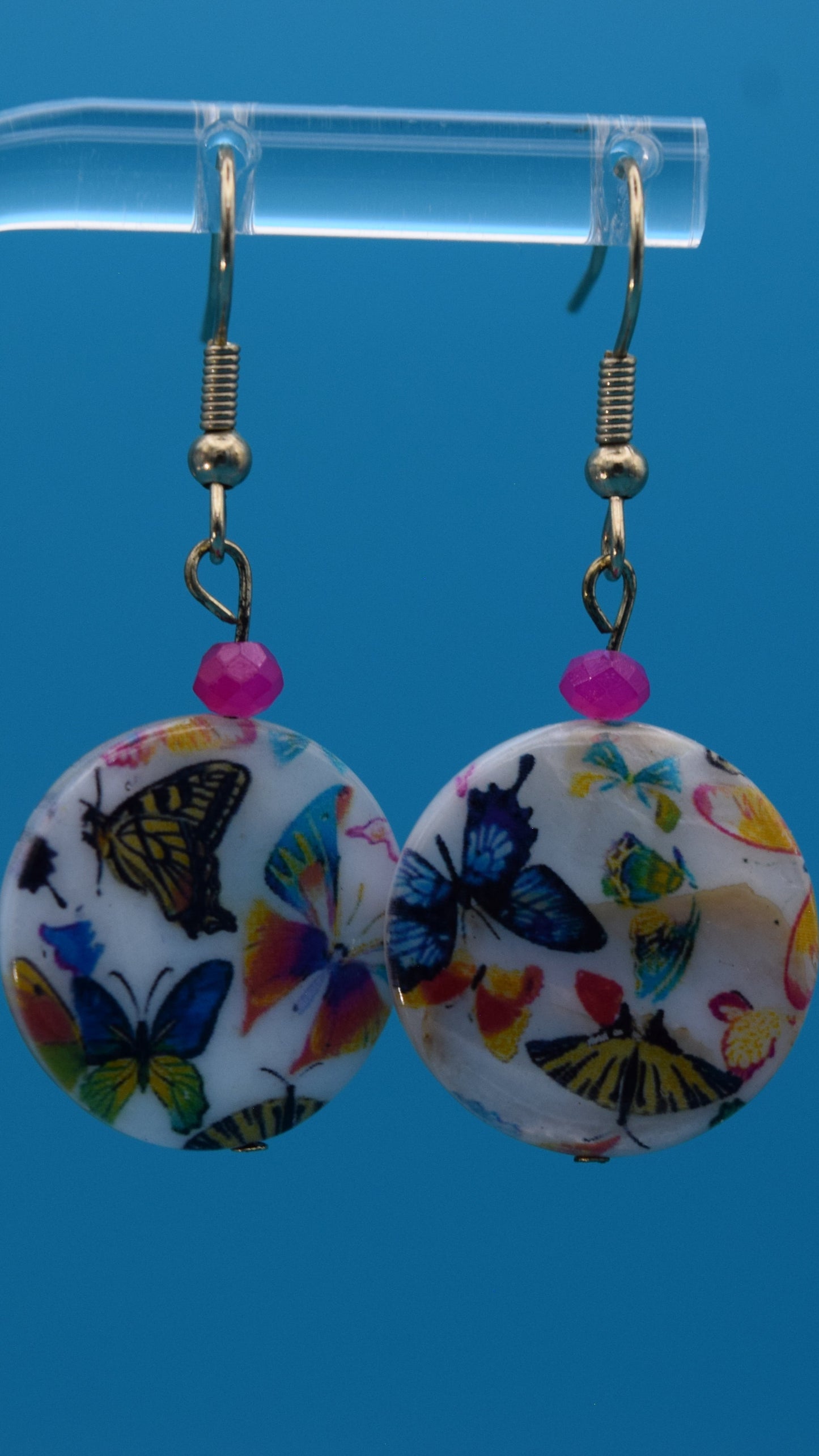 Colorful butterfly-patterned earrings Displayed on a clear stand against a teal background.