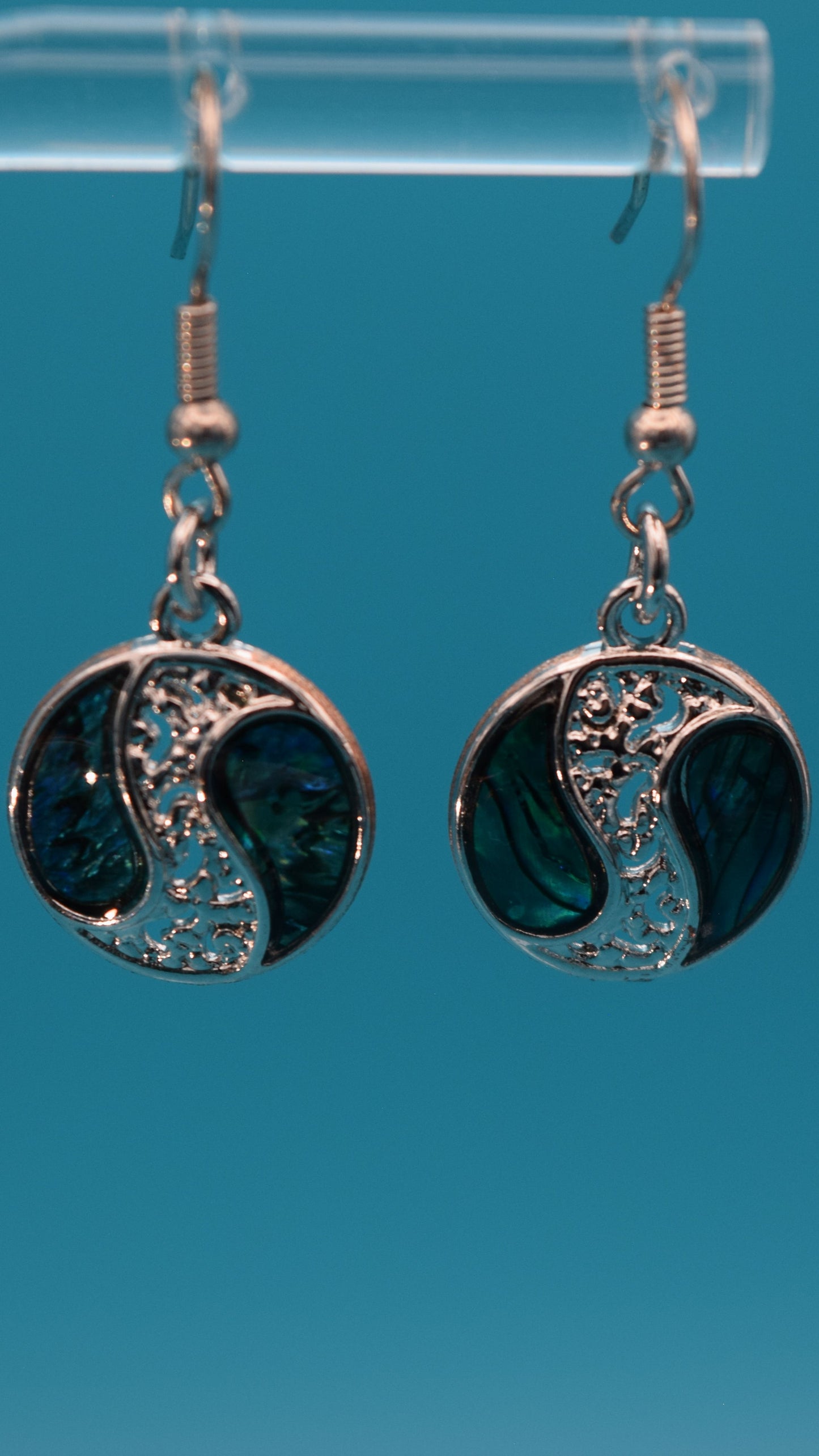 Silver yin yang style earrings dangling 3/4 inch, with iridescent blue, green pattern with black swirls on each side of a silver design. Displayed on a clear stand against a teal background.