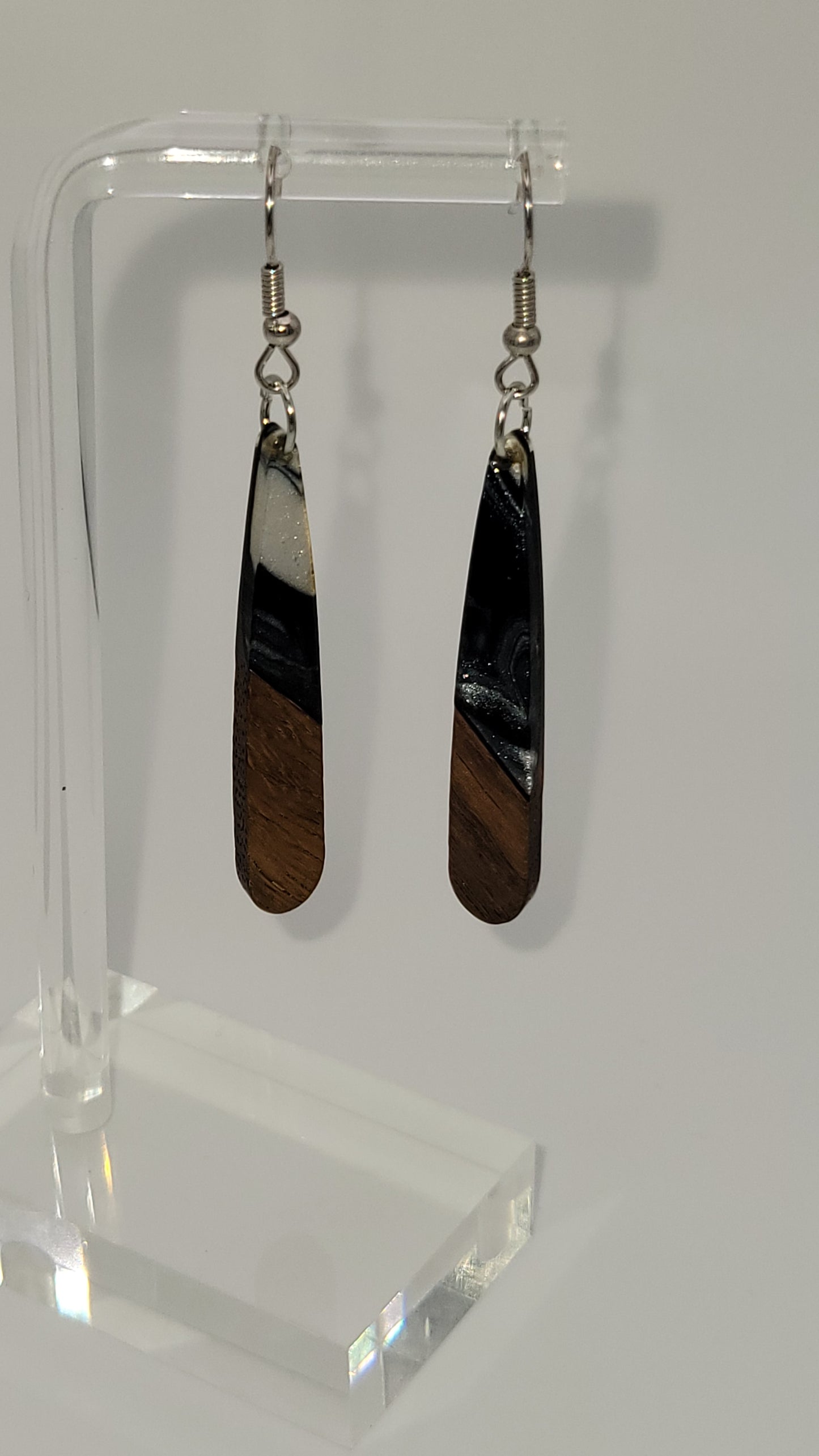 Long-gated tear shaped earrings made of wooden and epoxy earring a walnut colored wood.  Colored with a black, white sparkle epoxy. Displayed on a clear stand against a white background