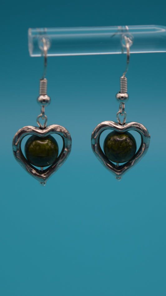 Silver heart-shaped dangle earrings hanging 1/2 inch, with a green bead  in the center of the heart. Displayed on a clear stand against a teal background.