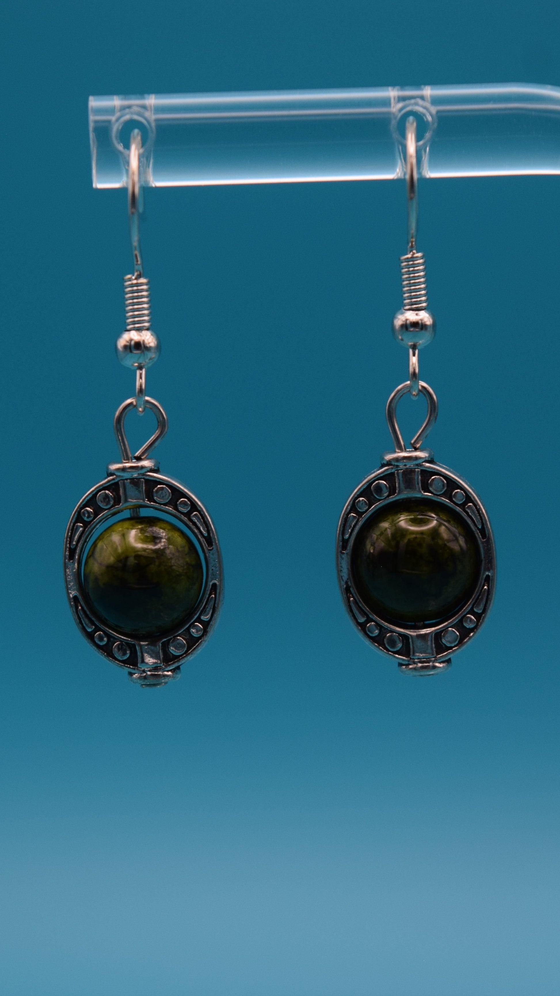 A pair of dangle earrings, each featuring a round, dark green bead set within an ornate, antique silver-colored metal bezel. The metalwork around the stone has a textured, appearance with small decorative dots around the edge. The bead itself is smooth and polished, with variations in its dark green color. This is displayed on a clear stand against a teal background.