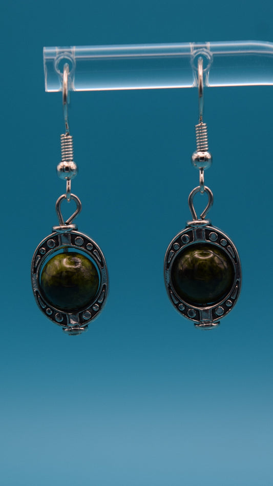 A pair of dangle earrings, each featuring a round, dark green bead set within an ornate, antique silver-colored metal bezel. The metalwork around the stone has a textured, appearance with small decorative dots around the edge. The bead itself is smooth and polished, with variations in its dark green color. This is displayed on a clear stand against a teal background.