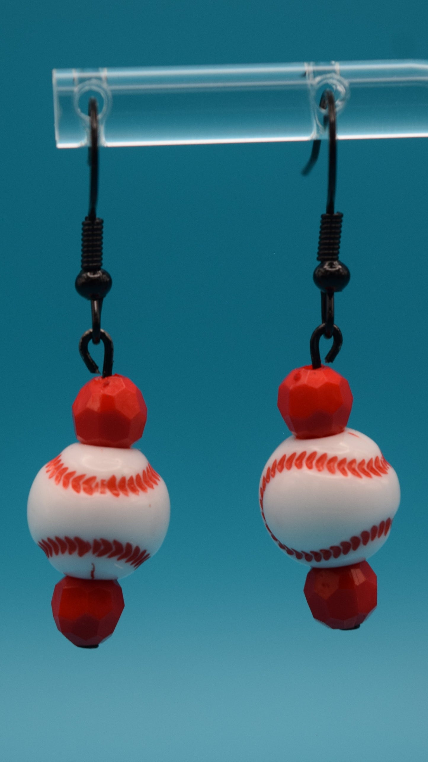 The top bead is a small, faceted bright red bead. the middle bead is a larger, round, smooth white bead that is painted to resemble a miniature baseball. It has the iconic red stitching pattern curving across its surface, just like a real baseball. The bottom bead is identical to the top bead. 