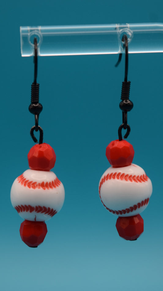 The top bead is a small, faceted bright red bead. the middle bead is a larger, round, smooth white bead that is painted to resemble a miniature baseball. It has the iconic red stitching pattern curving across its surface, just like a real baseball. The bottom bead is identical to the top bead. 