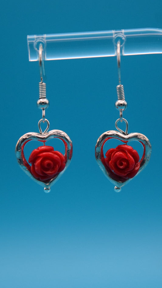 1/2 inch Silver Heart-shaped outline dangle earrings with red roses with a tiny silver ball at the bottom of the heart. Earrings are displayed on a clear stand against a teal background.