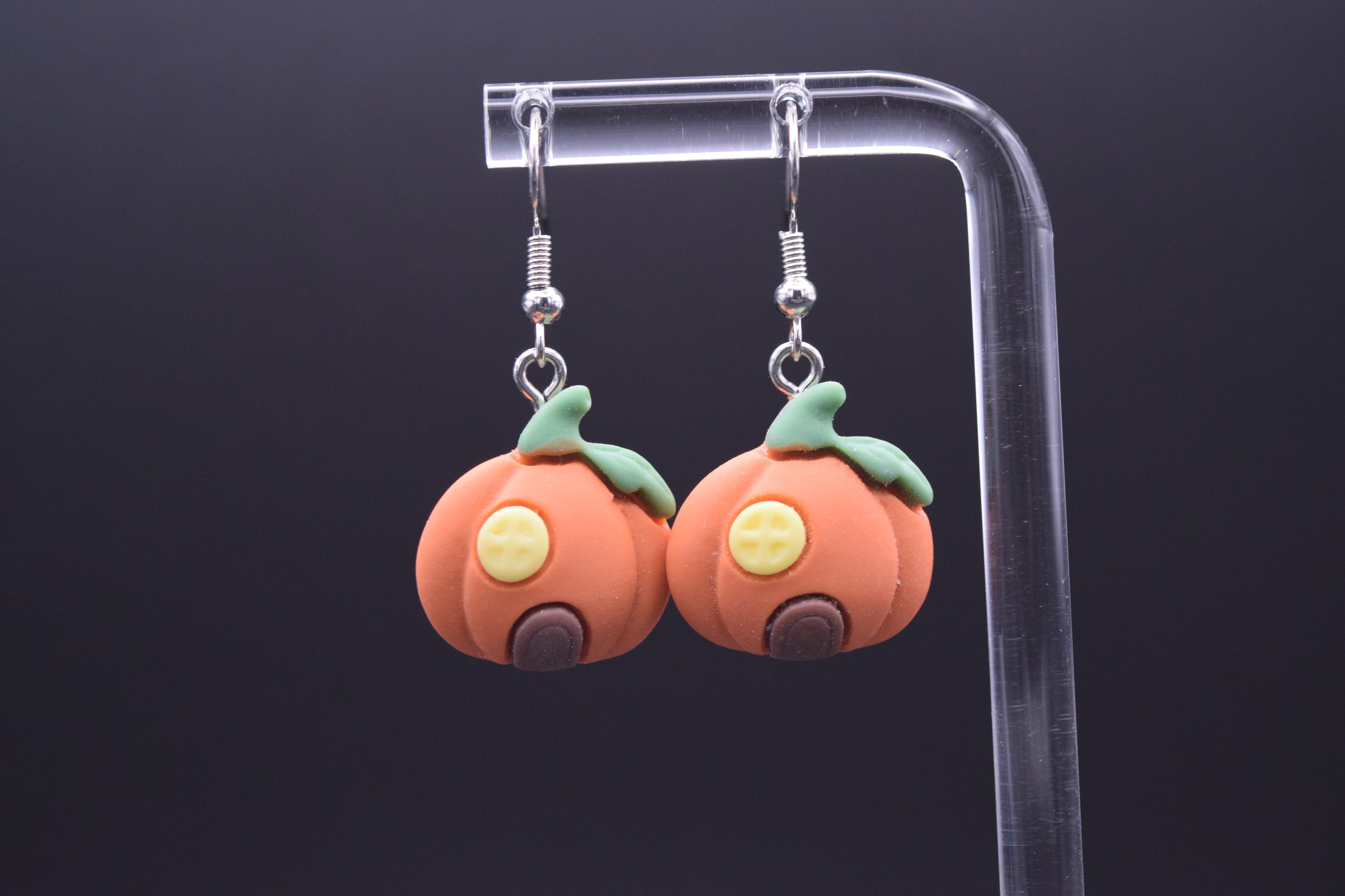 Each pumpkin features a circular yellow window with a  cross-hatch design, giving the impression of window panes. Below the window, there's a small, arched brown doorway, completing the "house" look. They are suspended from a clear, acrylic display stand against a black background. 