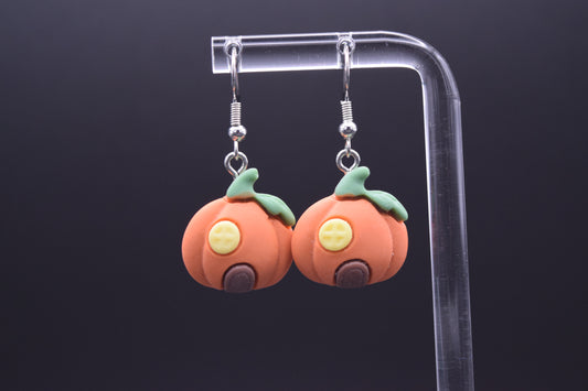 Each pumpkin features a circular yellow window with a  cross-hatch design, giving the impression of window panes. Below the window, there's a small, arched brown doorway, completing the "house" look. They are suspended from a clear, acrylic display stand against a black background. 