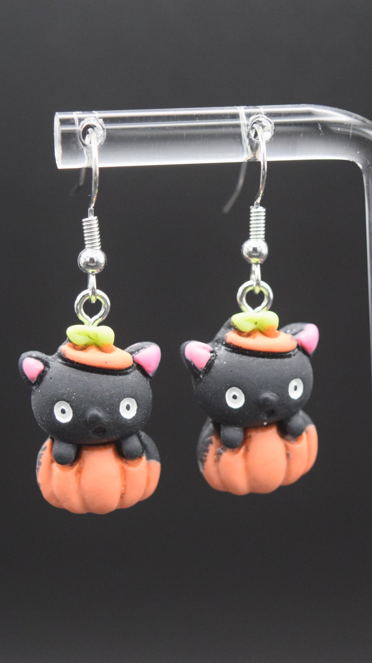 Each earring features a cute, stylized black cat head emerging from an orange pumpkin. The black cat heads have pink inner ears, white eyes, and a small black nose. On top of each cat's head, there's a tiny orange hat with a green stem, mimicking the top of a pumpkin. The orange pumpkin forms the body of the earring, with ridges indicating its shape. These are displayed on a clear acrylic stand with a black background.