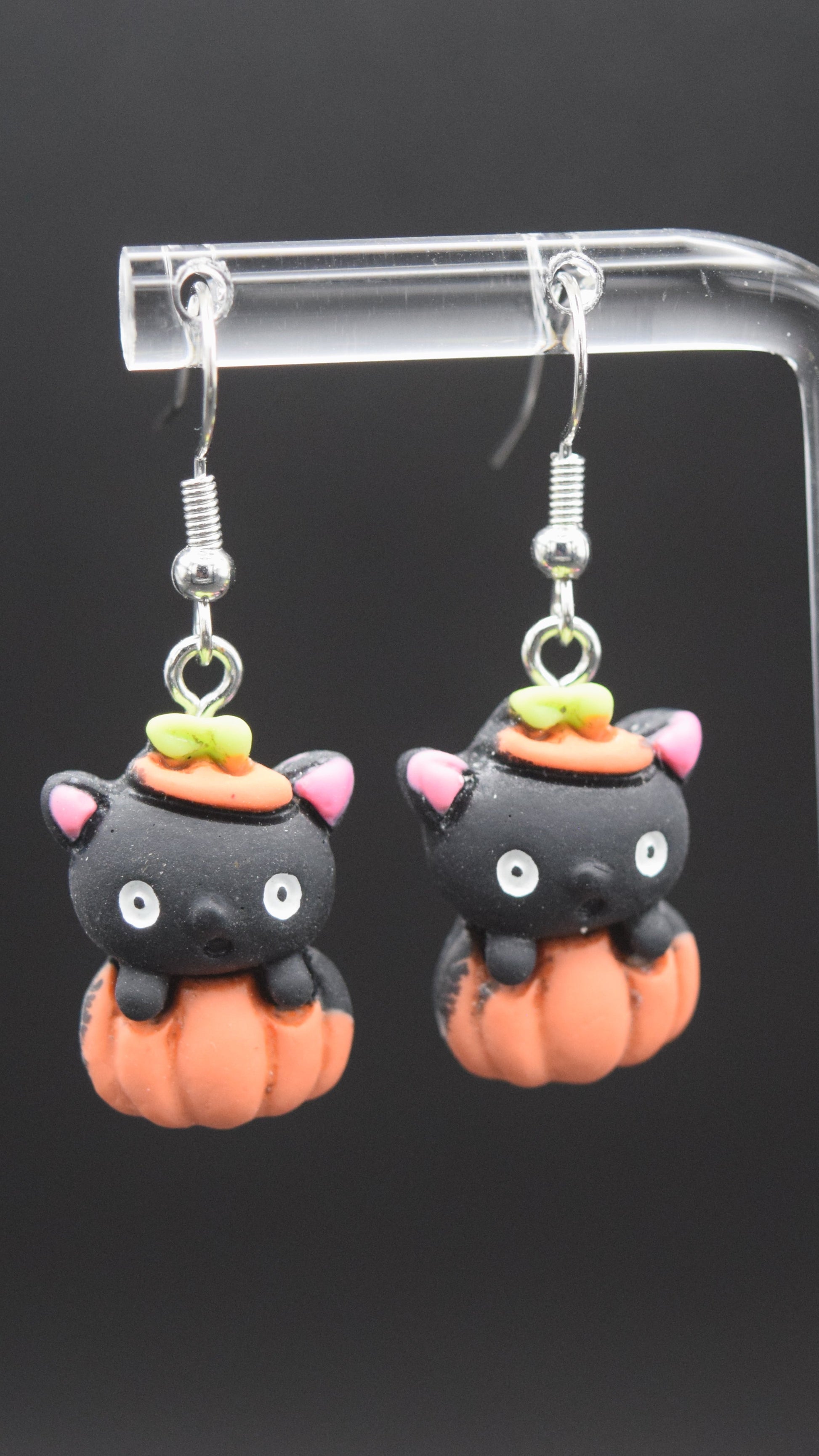 Each earring features a cute, stylized black cat head emerging from an orange pumpkin. The black cat heads have pink inner ears, white eyes, and a small black nose. On top of each cat's head, there's a tiny orange hat with a green stem, mimicking the top of a pumpkin. The orange pumpkin forms the body of the earring, with ridges indicating its shape. These are displayed on a clear acrylic stand with a black background.