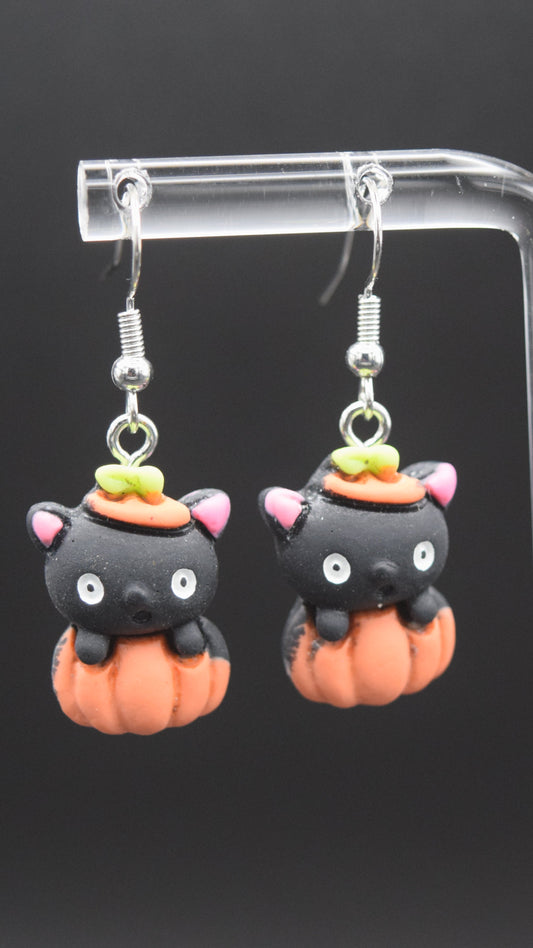 Each earring features a cute, stylized black cat head emerging from an orange pumpkin. The black cat heads have pink inner ears, white eyes, and a small black nose. On top of each cat's head, there's a tiny orange hat with a green stem, mimicking the top of a pumpkin. The orange pumpkin forms the body of the earring, with ridges indicating its shape. These are displayed on a clear acrylic stand with a black background.
