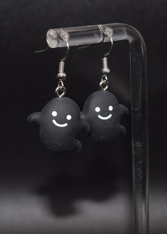 Black silicone ghost-shaped earrings with white smiley faces. Hanging off a on a clear stand against a black background