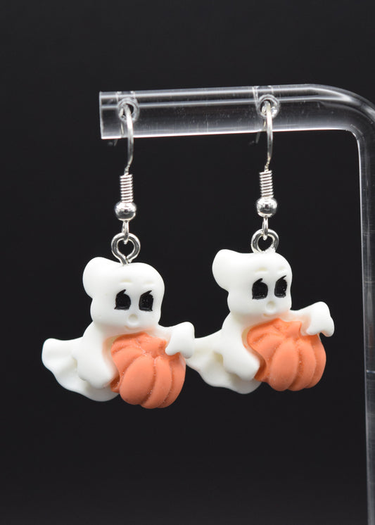 Silicone ghost-shaped earrings with white ghost, with black eyes, holding a orange pumpkins. Displayed on a clear stand against a black background