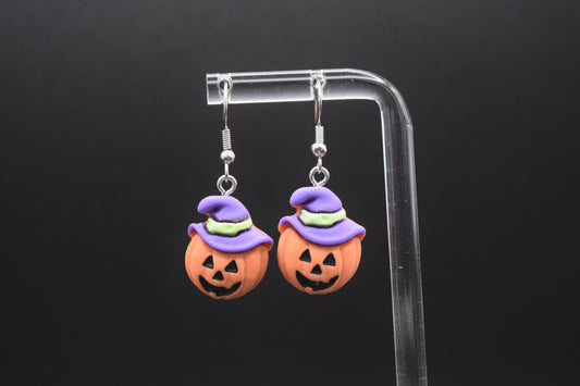 Each earring features a small, round, orange pumpkin with carved-out eyes and a jagged smile, giving it a classic jack-o'-lantern face. Perched on top of each pumpkin is a stylized witch's hat. The hats are primarily purple, with a light green band around the base. The top of the hat is pointed and slightly curved. Displayed on a clear acrylic stand against a black background.