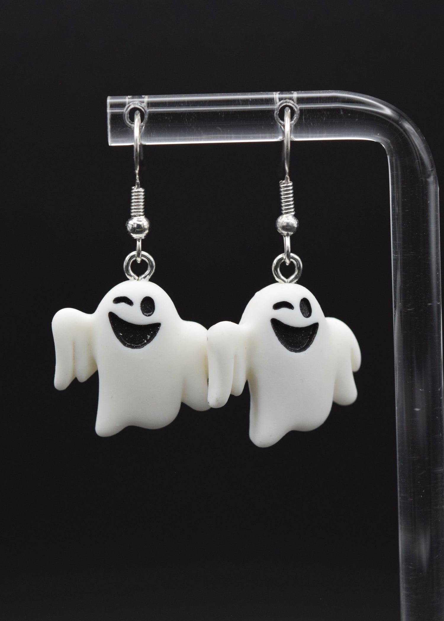 Silicone ghost-shaped earrings with a playful winking expression displayed on a clear stand against a black background
