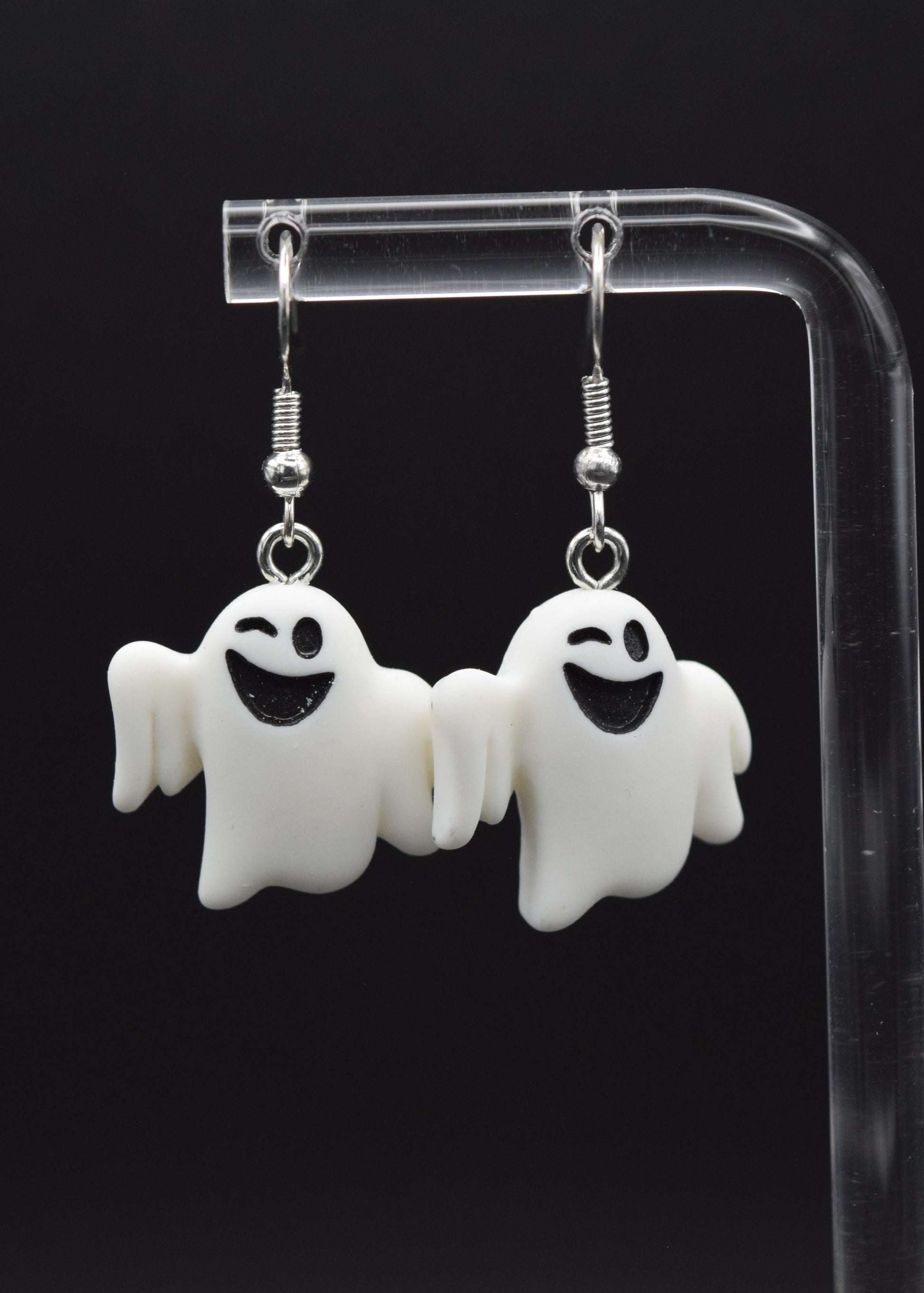 Silicone ghost-shaped earrings with a playful winking expression displayed on a clear stand against a black background