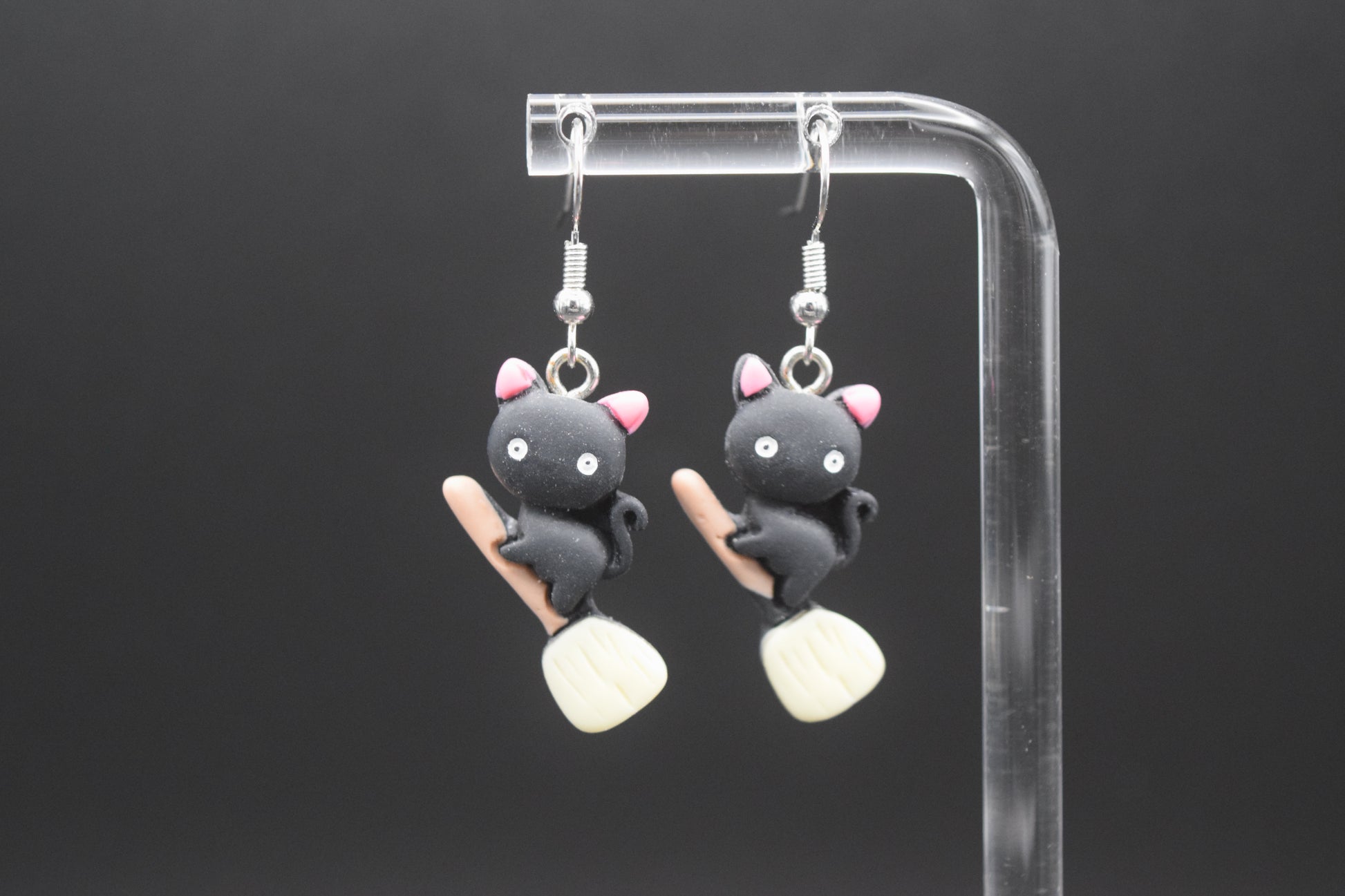 Each earring features a small, black, cat-like figure with pink inner ears, white eyes, and a tiny black nose. The cats are depicted in a stylized, slightly plump form. Below each cat, there's a miniature light brown broomstick with a pale yellow or off-white bristled end. The cats appear to be "riding" the broomsticks. They are displayed on a clear acrylic stand against a dark, featureless background. They are displayed on a clear acrylic stand against a dark background. 