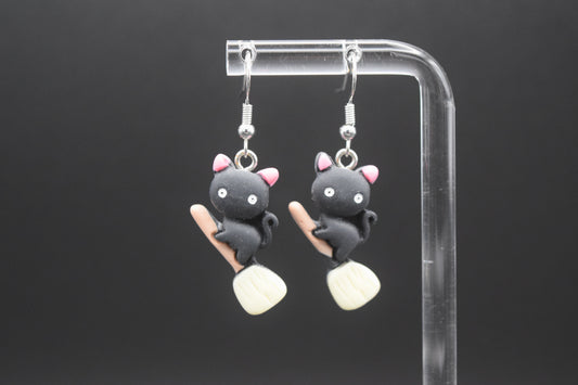 Each earring features a small, black, cat-like figure with pink inner ears, white eyes, and a tiny black nose. The cats are depicted in a stylized, slightly plump form. Below each cat, there's a miniature light brown broomstick with a pale yellow or off-white bristled end. The cats appear to be "riding" the broomsticks. They are displayed on a clear acrylic stand against a dark, featureless background. They are displayed on a clear acrylic stand against a dark background. 