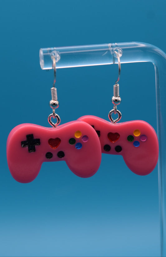 These are a playful and charming pair of dangle game controller earrings, made of colored pink epoxy, painted and topped with a clear coat. They're displayed hanging side-by-side on a clear acrylic stand, set against a vibrant, solid teal background.