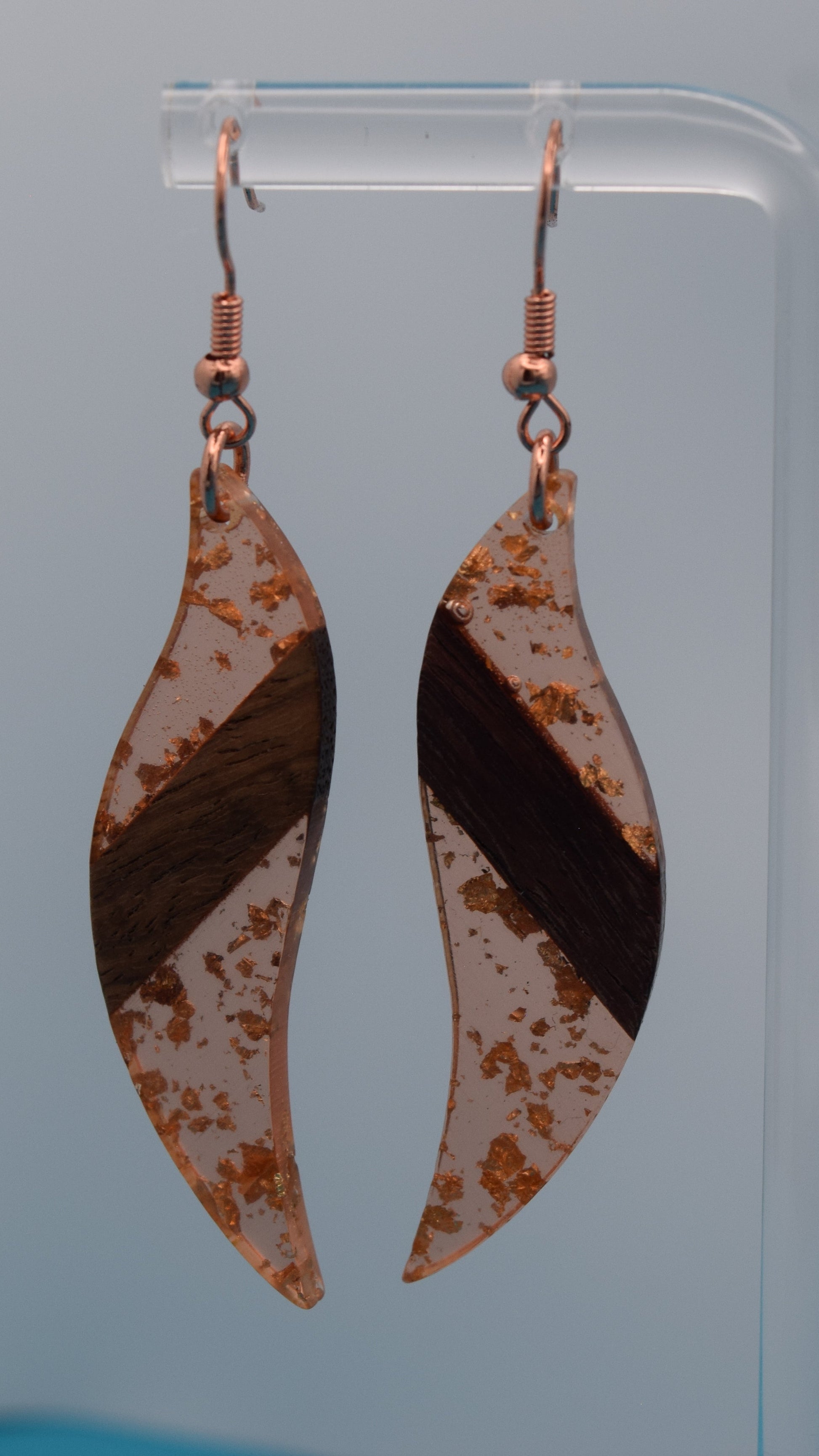 The earring is a translucent orange resin. This resin has a smooth, polished finish, allowing some light to pass through, giving it a luminous quality. Embedded within the resin are numerous small, irregular rose gold flakes with a walnut color piece of wood set in the middle of the earring. These flakes are scattered randomly, catching the light and adding a subtle, delicate shimmer and texture to the surface.