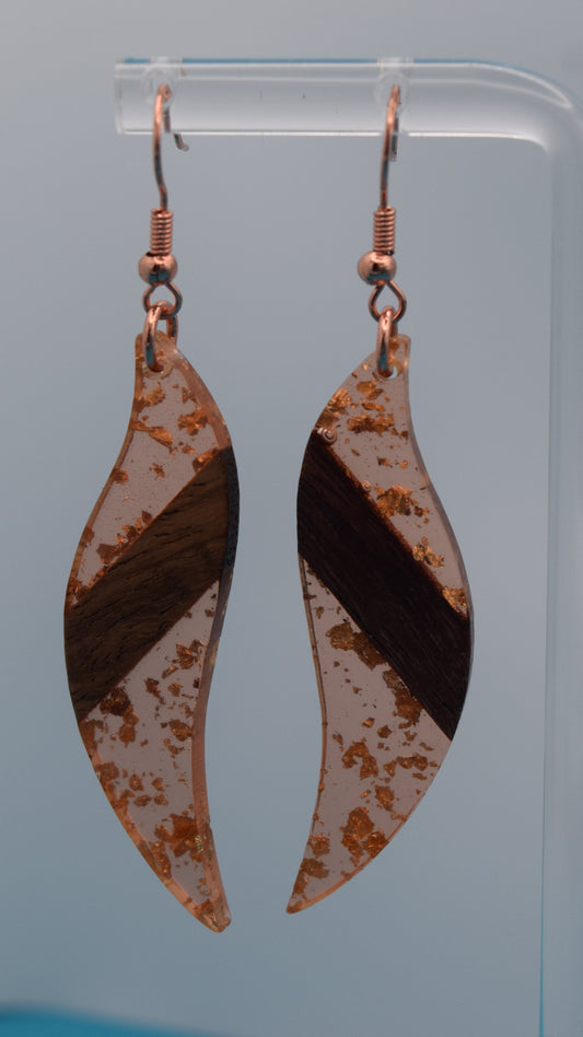 The earring is a translucent orange resin. This resin has a smooth, polished finish, allowing some light to pass through, giving it a luminous quality. Embedded within the resin are numerous small, irregular rose gold flakes with a walnut color piece of wood set in the middle of the earring. These flakes are scattered randomly, catching the light and adding a subtle, delicate shimmer and texture to the surface.